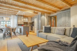 A modern kitchen with wooden beams, a dining area with chairs, a sofa with patterned cushions, and a wooden coffee table.