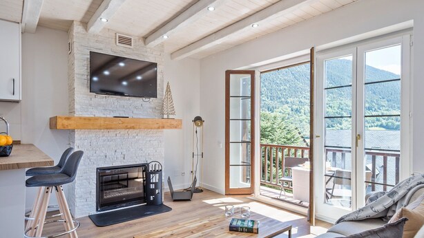 A modern living room with a fireplace, a TV mounted on a stone wall, and a large sliding glass door leading to a balcony with a mountain view. - Luderna - Apartamento con terraza Mirador Salardu (Naut Aran)