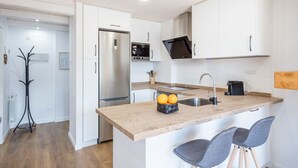 A modern kitchen with white cabinetry, a stainless steel refrigerator, a built-in oven, and a black induction cooktop. There are two bar stools at the island and a coat rack in the corner. - Luderna - Apartamento con terraza Mirador Salardu (Naut Aran)