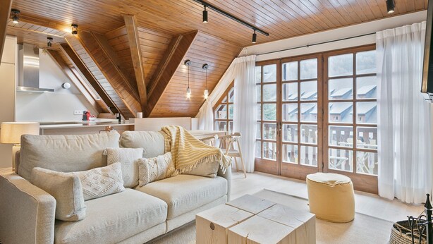 Ein gemütliches Wohnzimmer im Loft-Stil mit hohen Decken, Holzbalken, einem großen Fenster mit Aussicht, einer Couch mit Kissen und einem kleinen Holztisch.