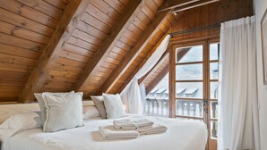 Ein Schlafzimmer mit Holzboden, einem großen Fenster mit Vorhängen, einem Bett mit Kissen und Handtüchern sowie einem gewebten Korb am Boden.