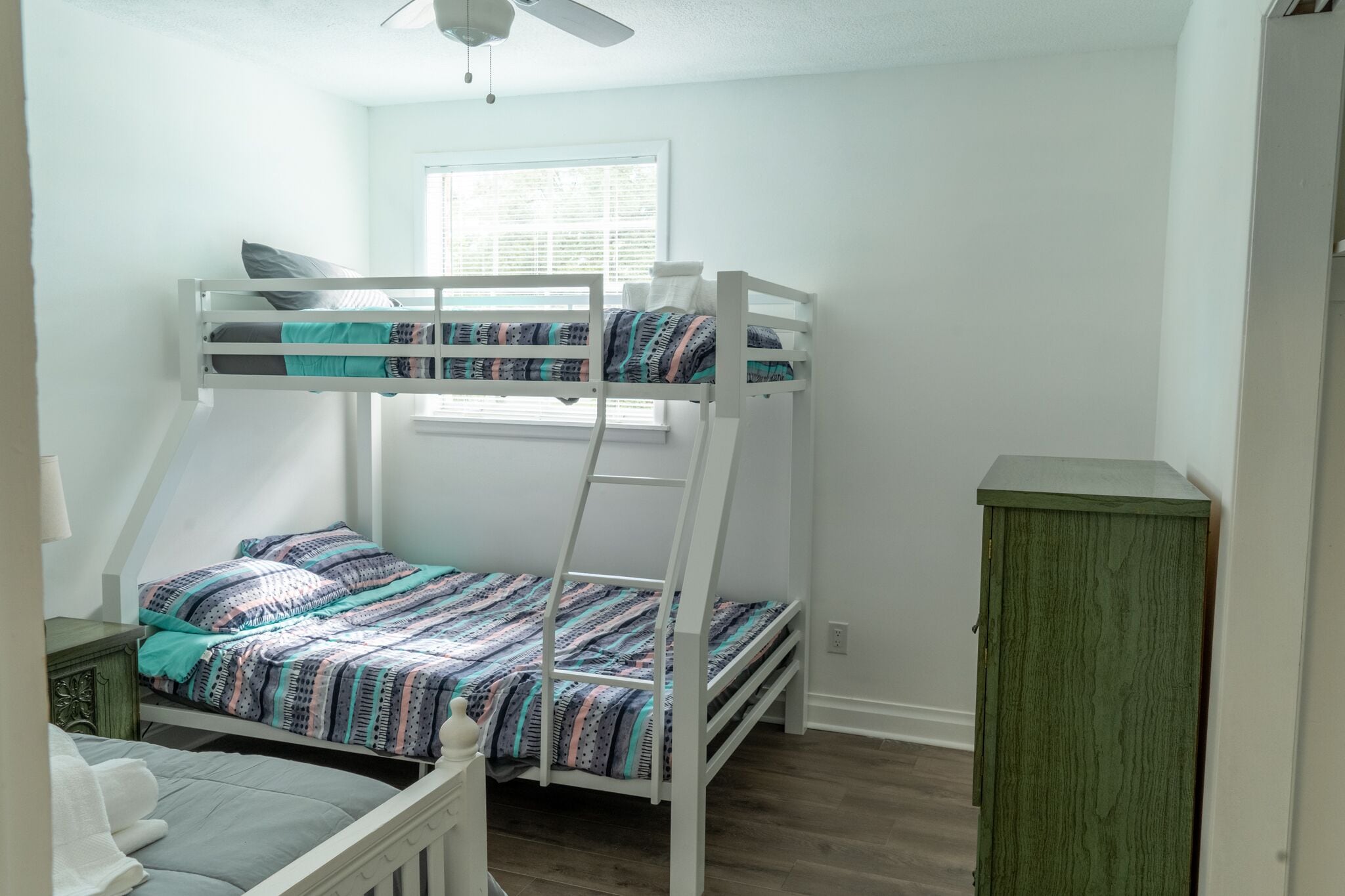 A bunk bed with a patterned bedspread, a small cabinet, and a window with blinds.