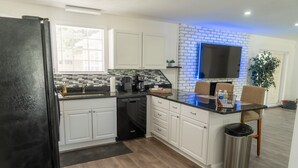Una cocina moderna con armarios blancos, un refrigerador negro, lavavajillas y cafetera. Hay una pared de ladrillo con un televisor montado sobre la encimera.