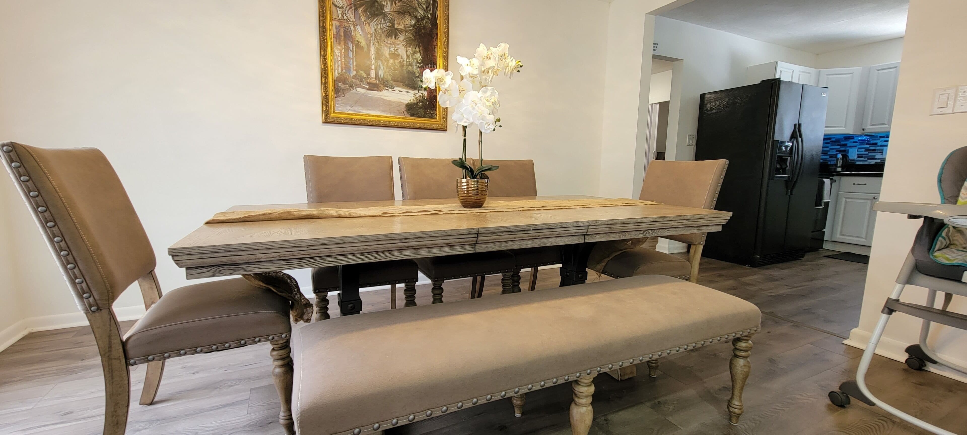 A dining area with a wooden table, cushioned chairs, and a bench. A vase with flowers is on the table.
