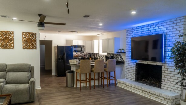 Una sala de estar moderna con chimenea, un comedor con taburetes de bar y una cocina con armarios blancos y un televisor grande.