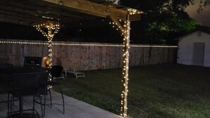 Un patio exterior con una pérgola de madera, luces colgantes, una parrilla y un cobertizo pequeño.