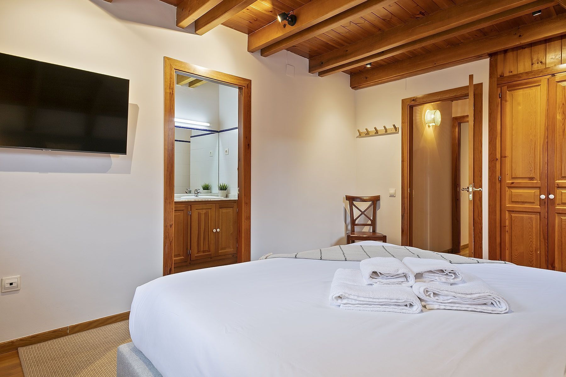 A bedroom with a bed, a mirror, a wooden wardrobe, and a television mounted on the wall.
