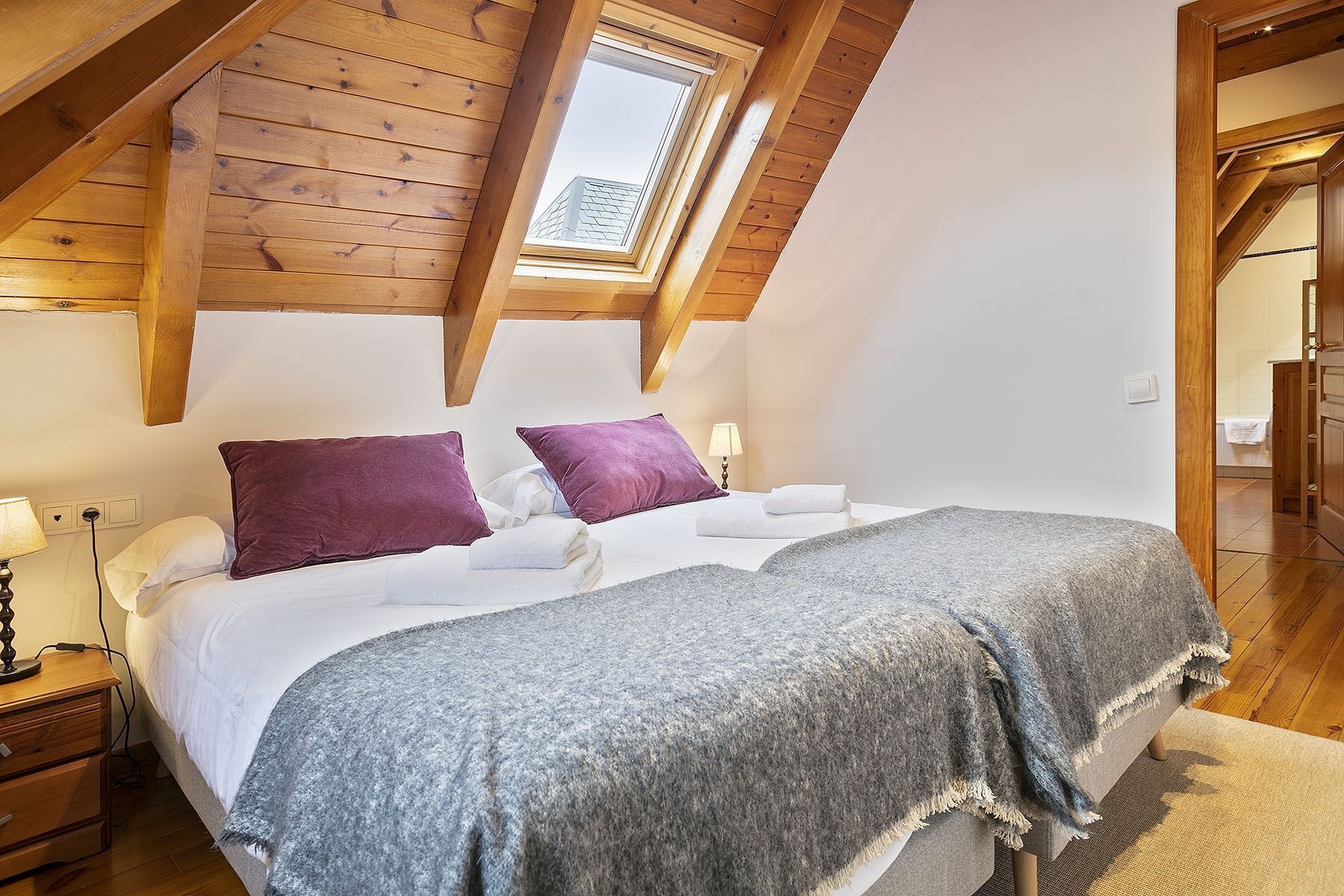 Une chambre avec un plafond en pente, des poutres en bois, un puits de lumière, un lit avec du linge de lit blanc, deux oreillers violets et une couverture grise.