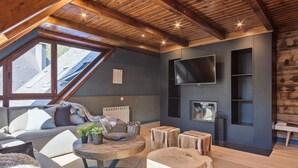 A cozy loft-style living room with a sloped ceiling, a large TV mounted on the wall, a fireplace, and a comfortable seating area with a sofa and coffee table. - Luderna - Tríplex Val de Ruda E3 der Humarau (Baqueira)