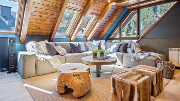 A cozy loft bedroom with a large window, wooden ceiling beams, a sectional sofa, a round coffee table, and a rustic wooden stool. - Luderna - Tríplex Val de Ruda E3 der Humarau (Baqueira)