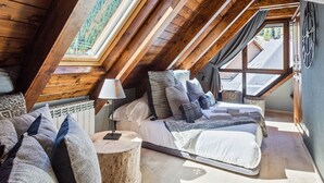 A cozy attic bedroom with a sloped ceiling, a large bed with multiple pillows, a bedside lamp, and a view of the outdoors through a window. - Luderna - Tríplex Val de Ruda E3 der Humarau (Baqueira)