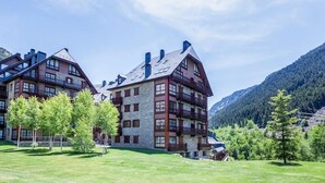 Um hotel de vários andares com fachada de pedra e varandas de madeira, rodeado por uma paisagem verde exuberante e montanhas ao fundo.