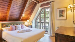 A bedroom with a large bed, wooden ceiling beams, a nightstand, a lamp, and a window with curtains.