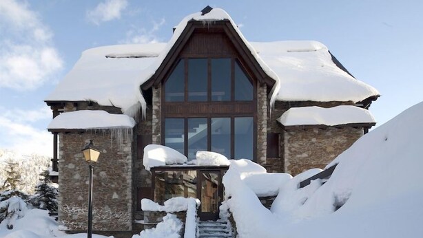 A stone building with a steep roof, large windows, and a wooden door. Snow covers the roof and ground.