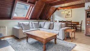 A loft-style living room with a wooden ceiling, a grey sectional sofa, a wooden coffee table, and a dining area with wooden chairs and a table.