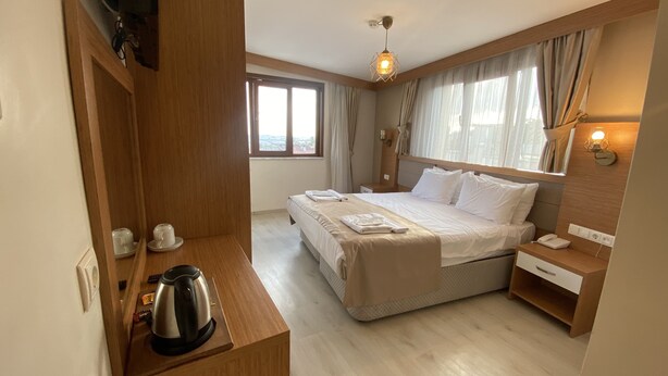 Une chambre d’hôtel comprenant un lit, une table de chevet, une bouilloire, un miroir, une fenêtre avec des rideaux et un luminaire fixé au mur.