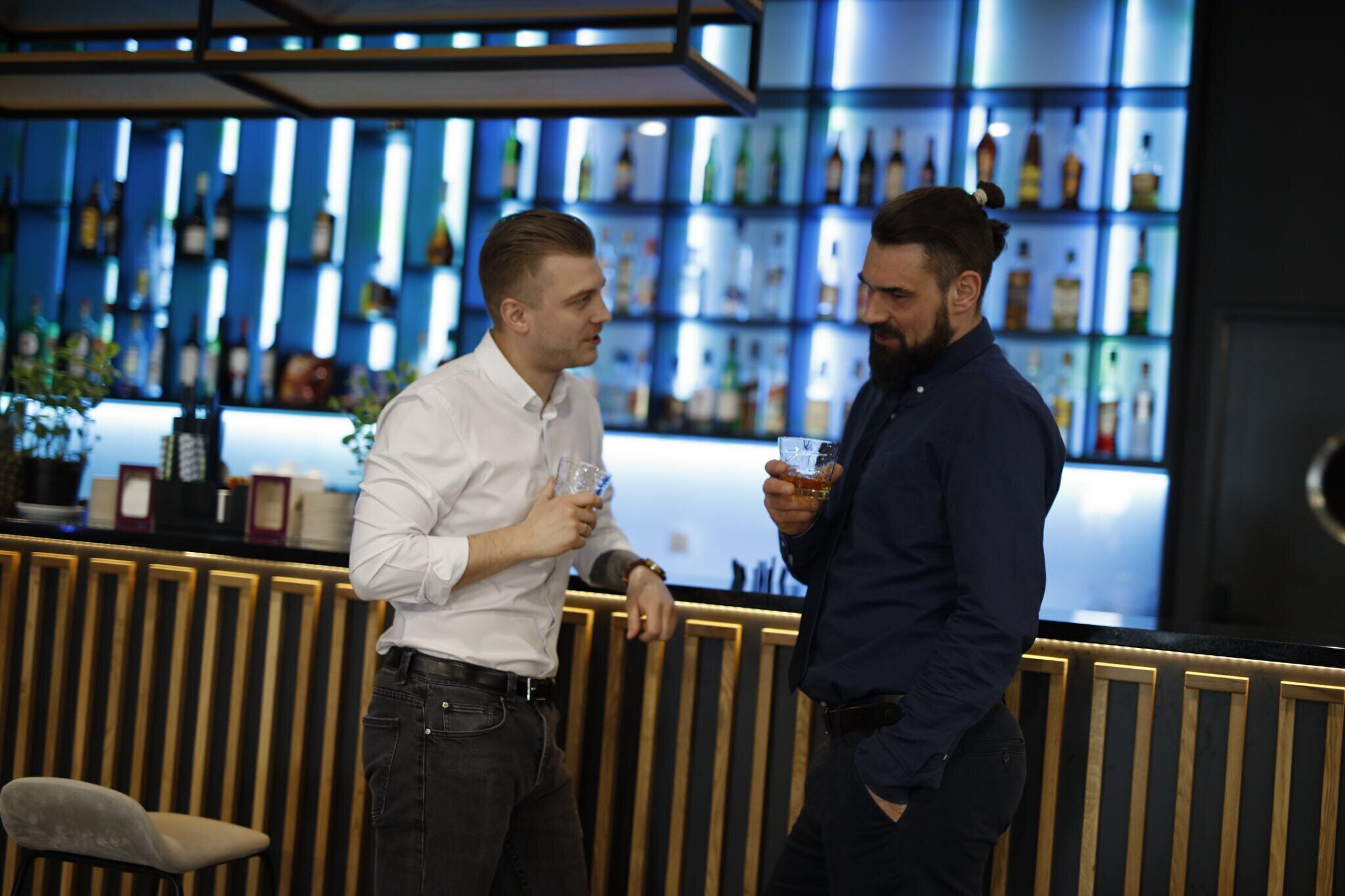 Due uomini chiacchierano in un bar dal design moderno, con un bancone e scaffali pieni di bottiglie.