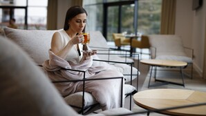 A person sitting on a sofa in a modern, well-lit room with large windows, a small round table, and a chair. - Hotel Robert's Port (Mikolajki)