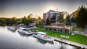 A waterfront hotel with a docked boat, a grassy area with lounge chairs, and a modern building with large windows. - Hotel Robert's Port (Mikolajki)