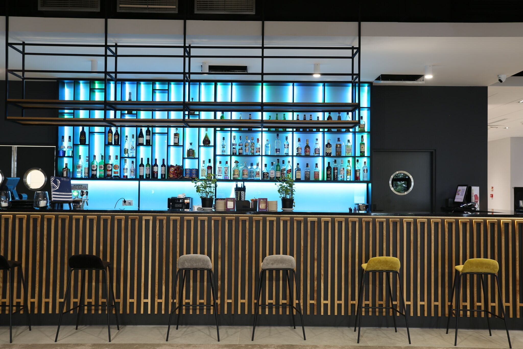 A bar area with a blue-lit shelf displaying various bottles, a reception desk, and four bar stools.