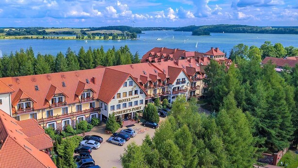 A hotel with red-tiled roofs, multiple stories, and a view of a lake with sailboats. - Hotel Robert's Port (Mikolajki)