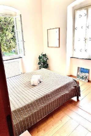 Un dormitorio con cama, ventana, radiador, planta y un cuadro en la pared.