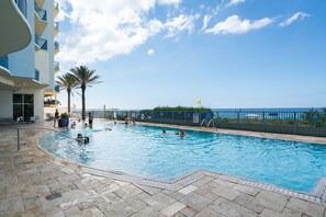 Une piscine où des gens nagent et s’amusent, entourée d’un bâtiment doté de balcons et offrant une vue sur la mer.