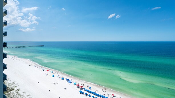 Een strand met helder turquoise water, een pier in de verte en parasols langs de kust.