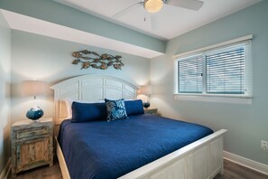 Une chambre à coucher comprenant un lit, des tables de chevet, un ventilateur de plafond et une fenêtre avec des stores.