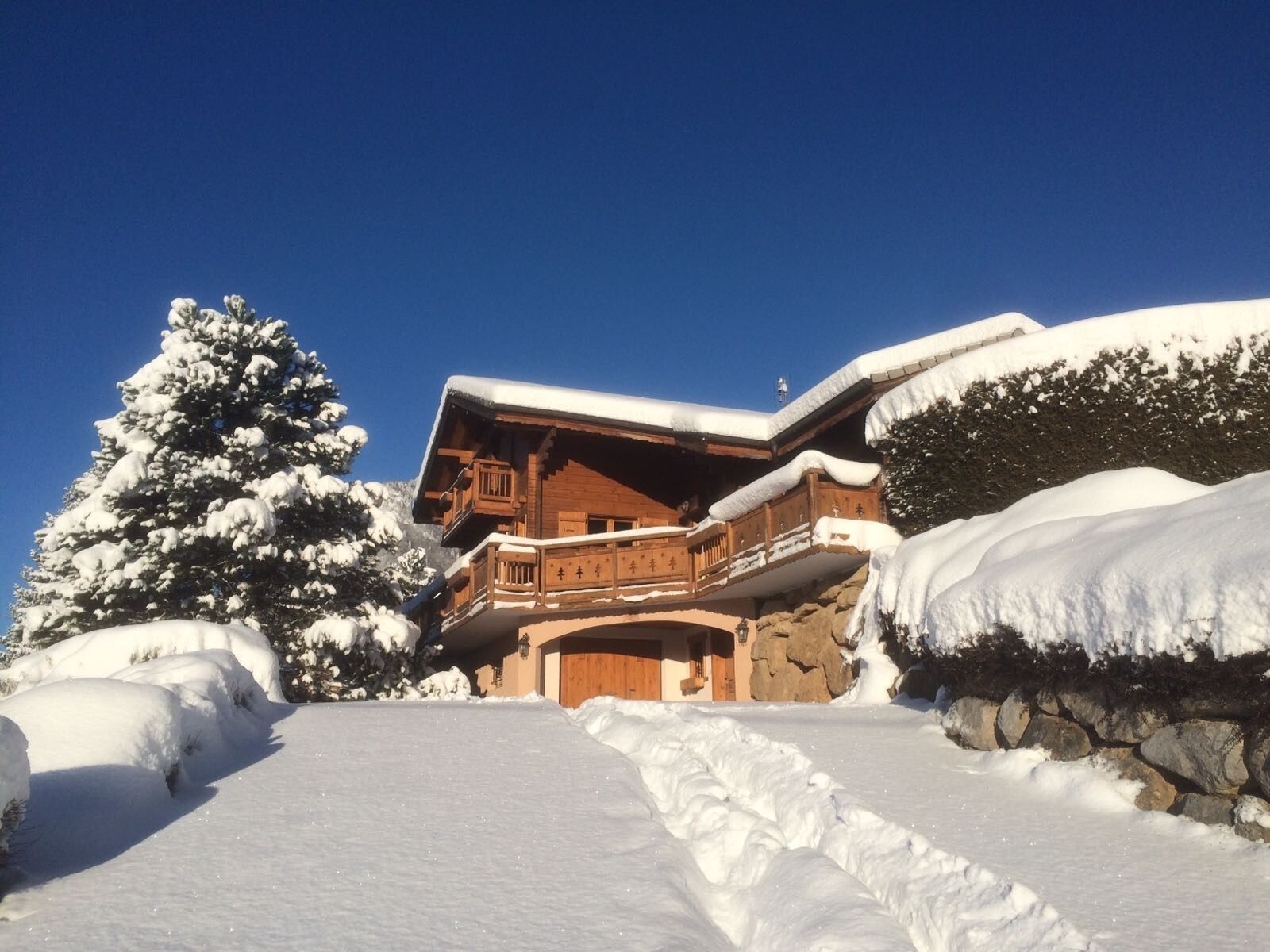 Un chalet en bois doté d’un balcon, entouré d’arbres couverts de neige et d’un mur de pierre.