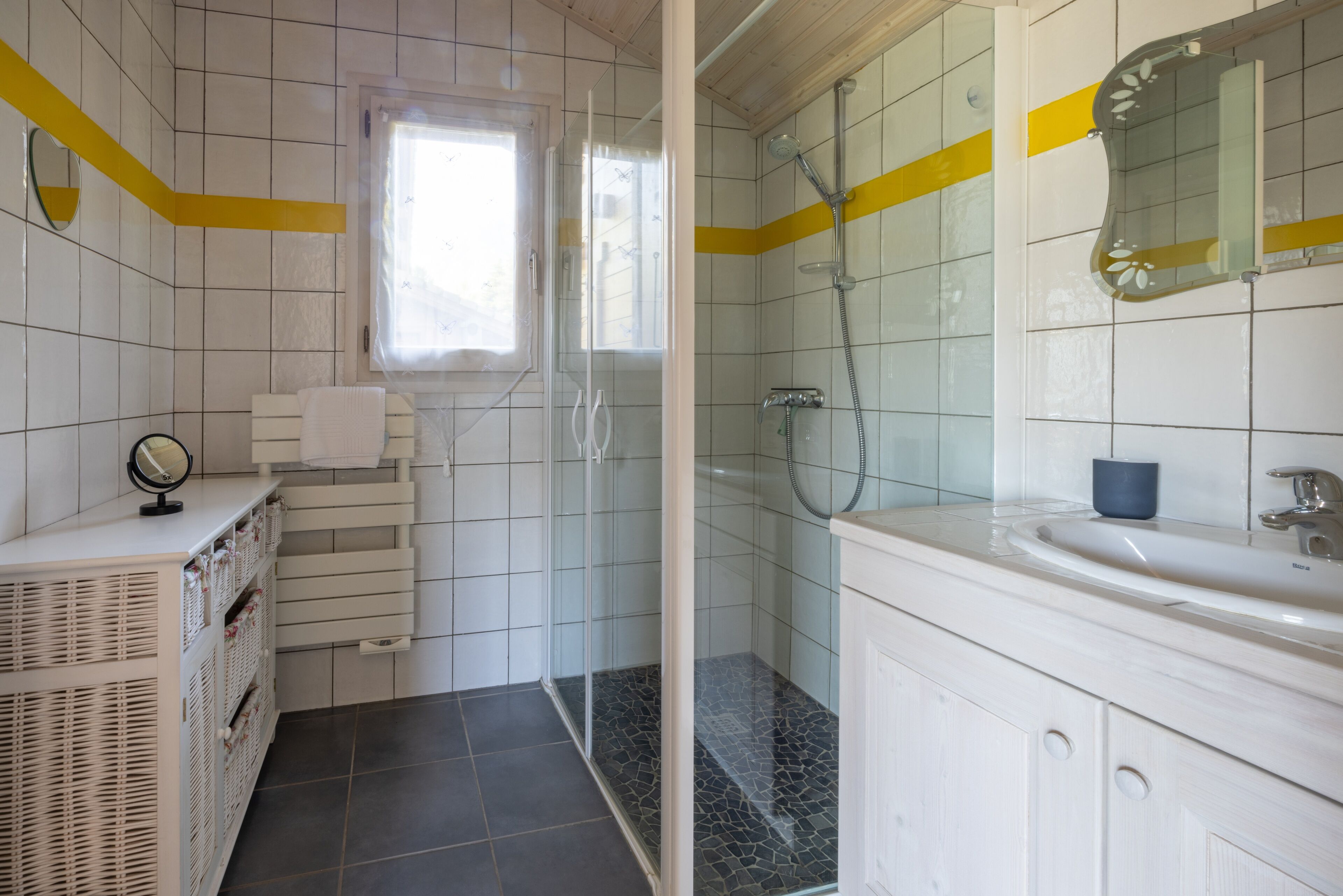 Un baño con paredes de azulejos blancos, una ducha con puerta de vidrio, un lavamanos con espejo y un ventanal que proporciona luz natural.