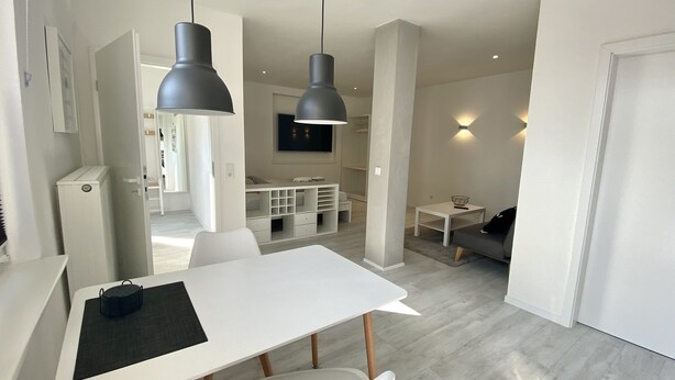 A modern, well-lit dining area with a white table and chairs, pendant lights, and a visible kitchen in the background. - Apartment Schloofschdubb in Fuldatal bei Kassel (Fuldatal)