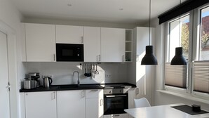 A modern kitchen with white cabinets, a black microwave, and a stove. There is a dining table with chairs and a window with blinds. - Apartment Schloofschdubb in Fuldatal bei Kassel (Fuldatal)