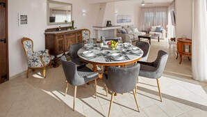 Un coin repas avec une table ronde dressée pour six personnes, entourée de six chaises grises à revêtement en tissu et à pieds en bois. Un buffet en bois, agrémenté d’un miroir et d’objets décoratifs, est adossé au mur.