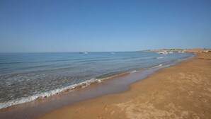 Une plage de sable fin, avec de douces vagues, des bateaux à l’horizon et un ciel dégagé.