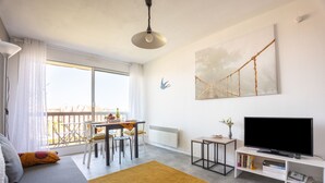 A modern living room with a sofa, a TV, a dining table with chairs, a small side table, and a large window with curtains. - Sunan - Appartement avec balcon (Courseulles-sur-Mer)