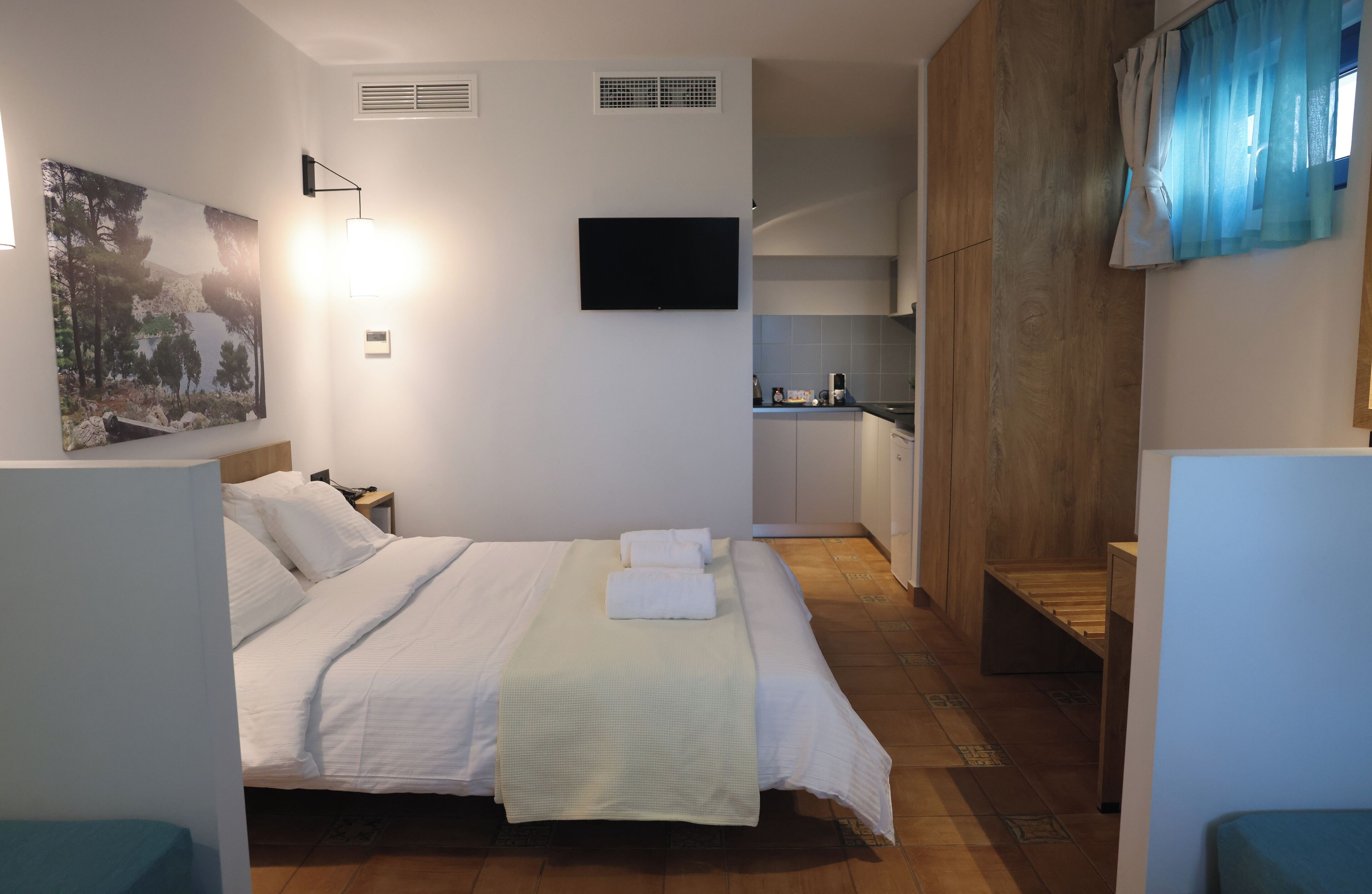 A hotel room with a bed, a flat-screen TV mounted on the wall, a small kitchen area, and a bathroom visible through an open door.