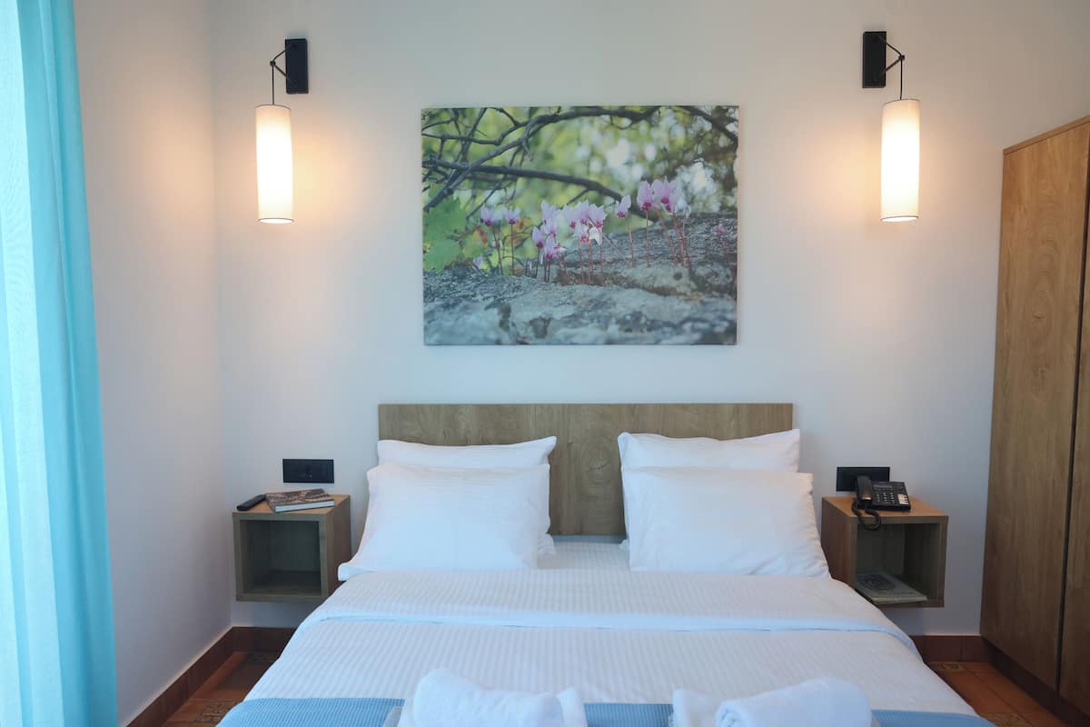 A neatly made bed with white linens, two bedside tables with lamps, a wooden wardrobe, and a painting of flowers on the wall.