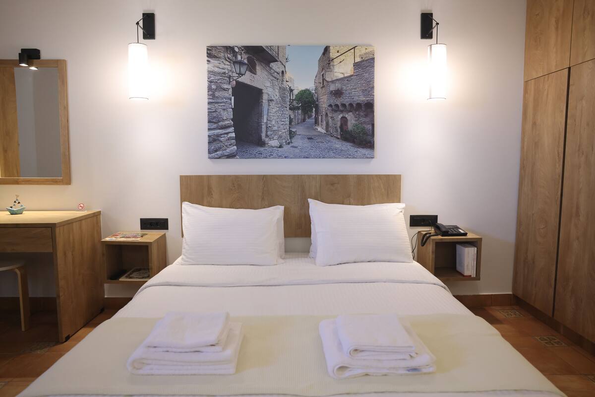 A hotel room with a wooden headboard, a bed with white linens, two bedside tables with lamps, a wooden desk, and a wall-mounted picture of a street scene.