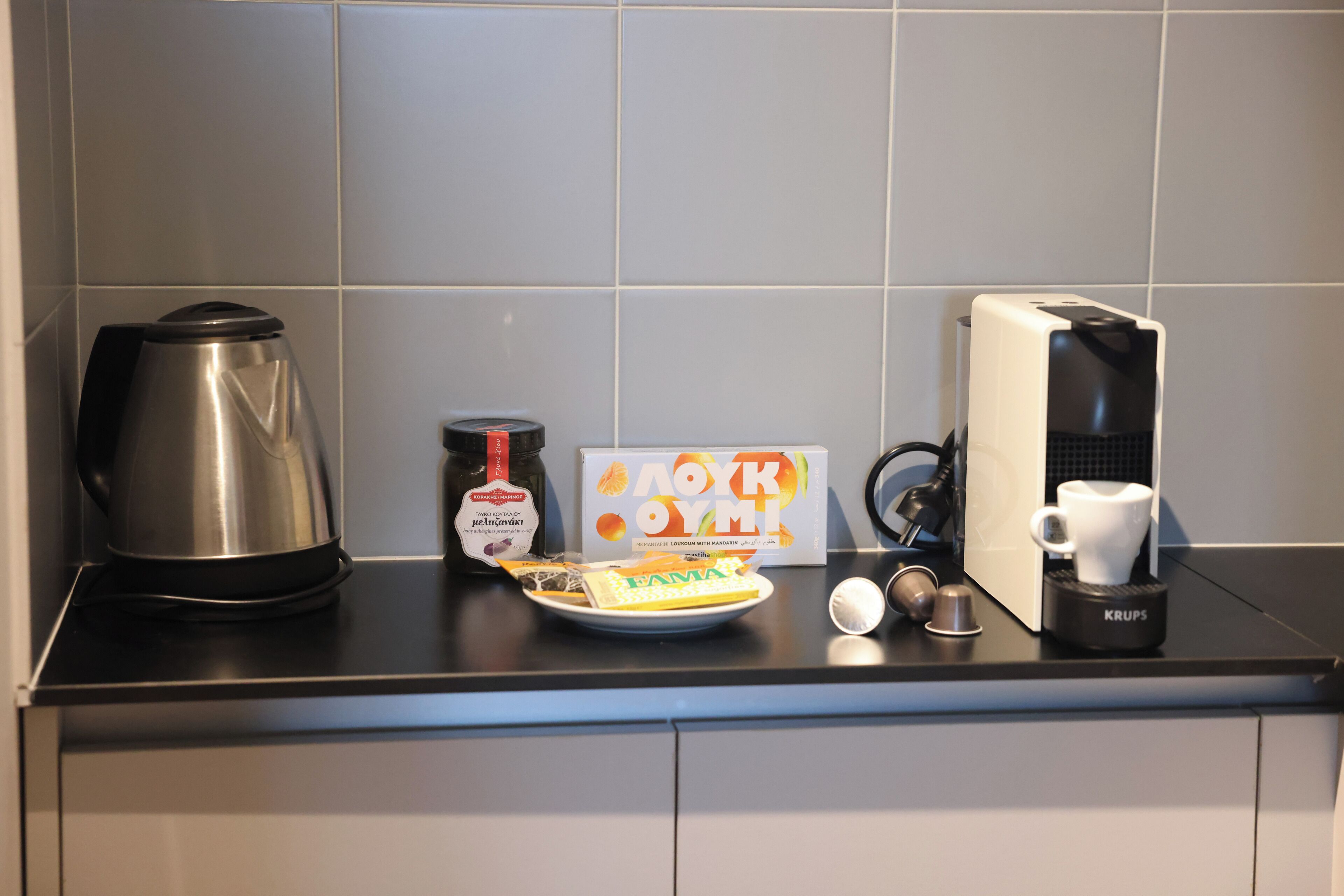 A kitchen counter with a coffee maker, a kettle, a plate with food, and a box of coffee pods.