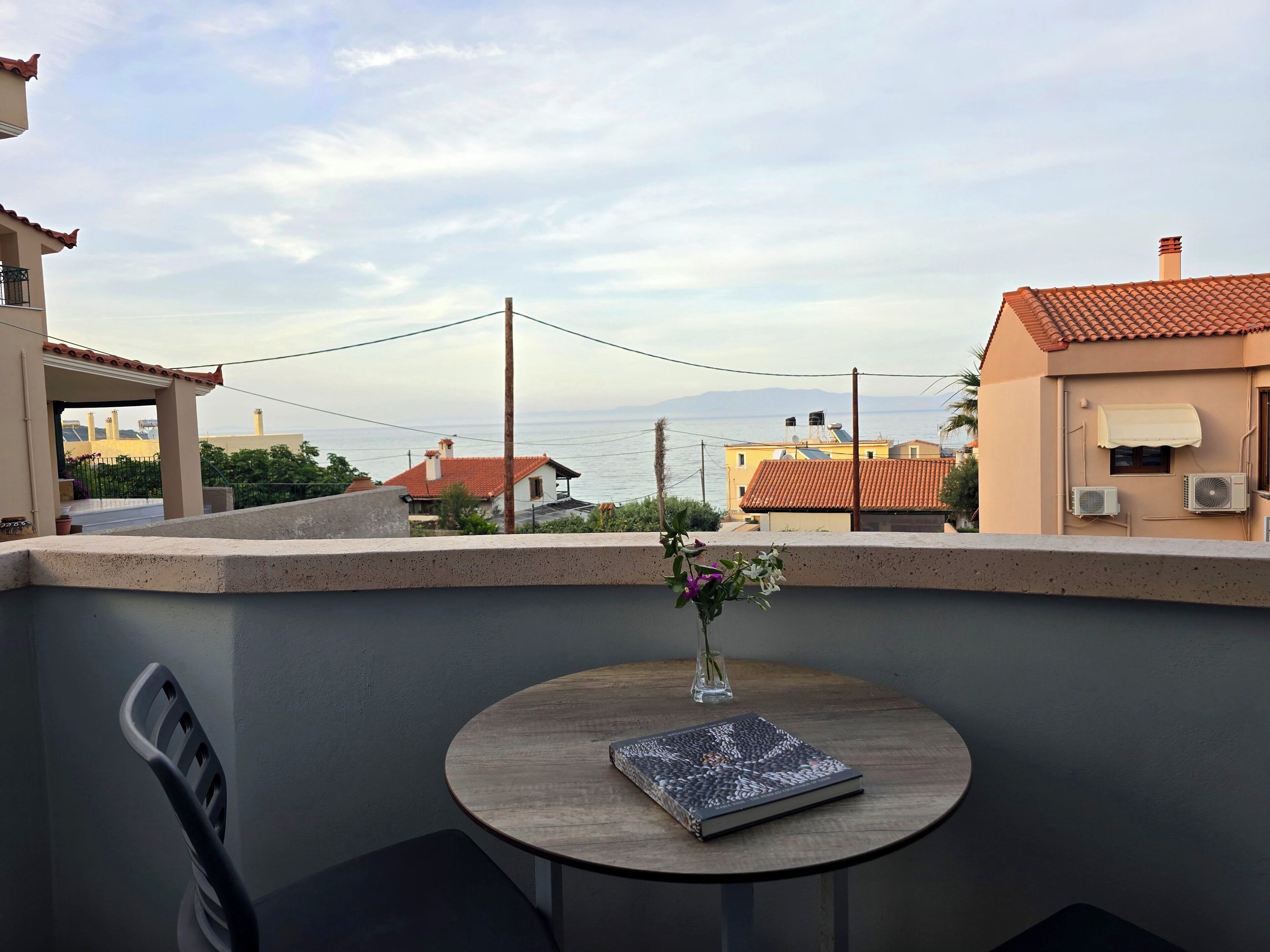 Un balcon avec une table, un vase rempli de fleurs et une vue sur la mer et les bâtiments.