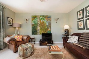 A living room with a brown leather sofa, a wooden coffee table with chess pieces, a television on a wooden stand, and a large abstract painting on the wall.