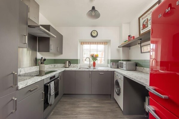 A compact kitchen with grey cabinets, a red refrigerator, a washing machine, and a clock on the wall.