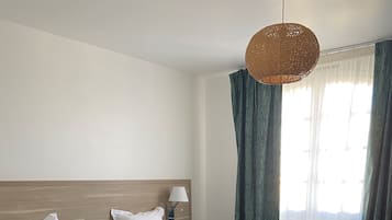 Habitación de hotel con dos camas, cada una con ropa blanca y almohadones verdes. Cuenta con un cabecero de madera, una mesita de noche con lámpara, y un ventanal con cortinas.