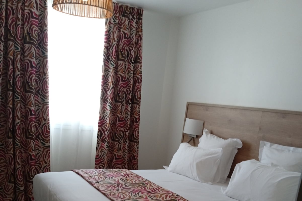 Una habitación de hotel con una cama grande, cabecera de madera, ropa de cama blanca y cortinas estampadas.