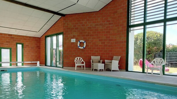 Une piscine intérieure avec un mur en briques, de grandes fenêtres et des chaises longues blanches.