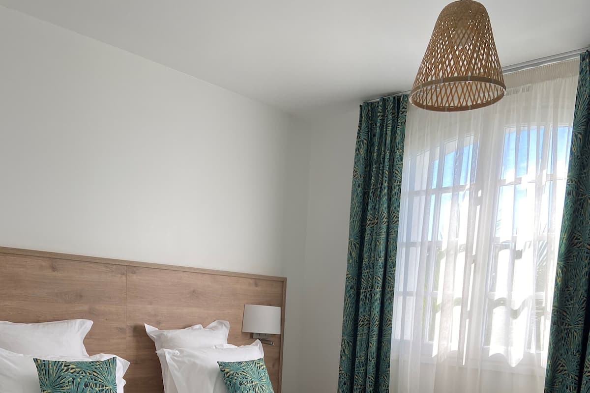 A neatly made bed with white linens and a green leaf-patterned throw, a wooden headboard, and a hanging light fixture.