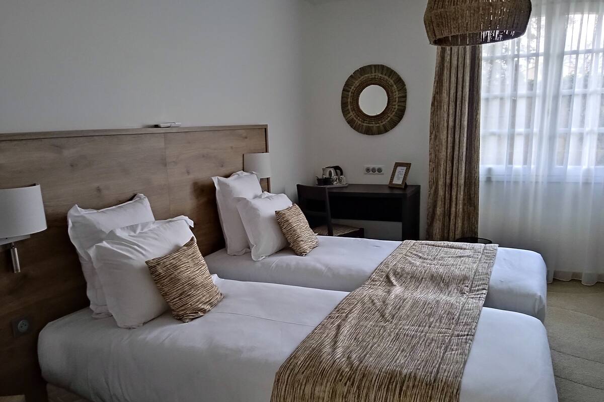 A hotel room with two beds, a wooden headboard, a desk with a lamp, a mirror, and a window with curtains.