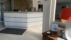 Un espace d’accueil avec un comptoir de réception blanc, une petite table en bois avec une lampe et un grand miroir qui reflète la pièce.