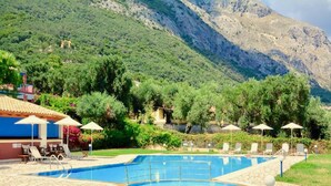 En swimmingpool med liggestole og parasoller, omgivet af grøn vegetation og bjerge.